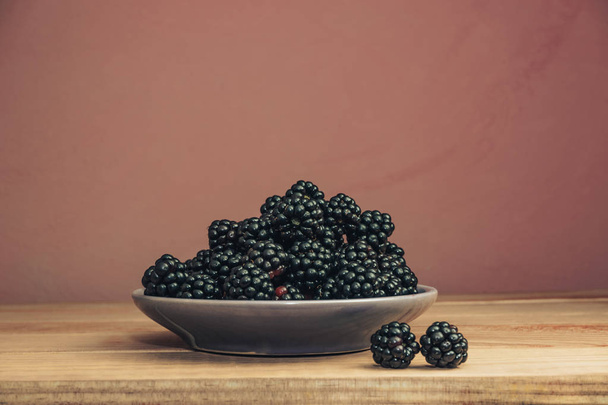 Fresh blackberry in bowl on a brown wooden table and dark-red wall background. - Valokuva, kuva