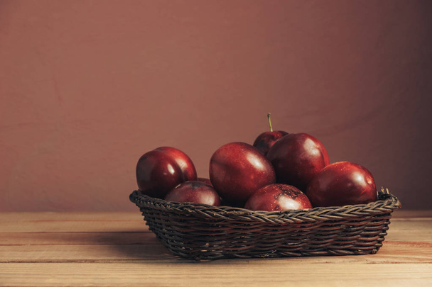 Fresh red plums in basket on a brown table. Beautiful dark red wall background. - Fotó, kép