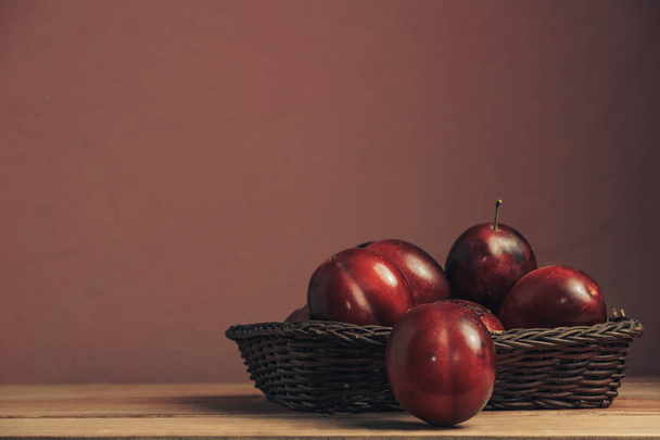 Fresh red plums in basket on a brown table. Beautiful dark red wall background. - Photo, Image