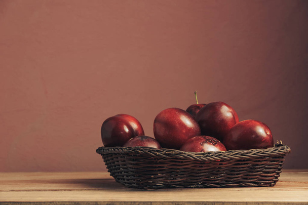 Fresh red plums in basket on a brown table. Beautiful dark red wall background. - Фото, изображение