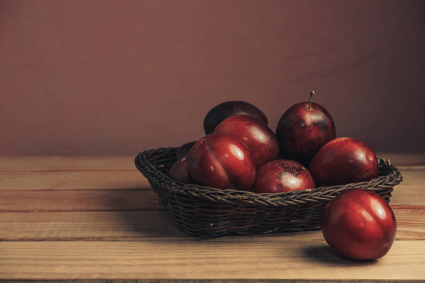 Fresh red plums in basket on a brown table. Beautiful dark red wall background. - Фото, изображение