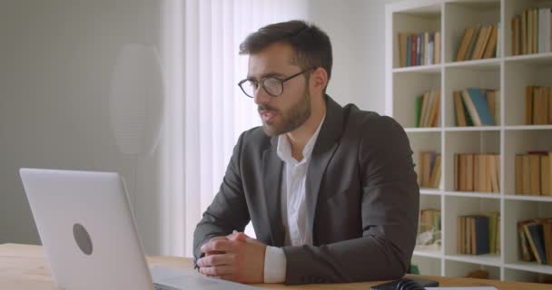 Closeup portrait of adult handsome bearded caucasian businessman in glasses having a video call on the laptop in the office indoors with bookshelves on the background - Footage, Video