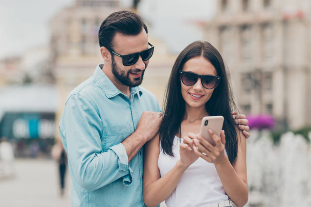 Portrait of excited married people have free time hold hand mobile device search look read eyewear eyeglasses brunettes hairstyle hug embrace touch shoulders shirt blue denim jeans outfit town walk - Photo, image