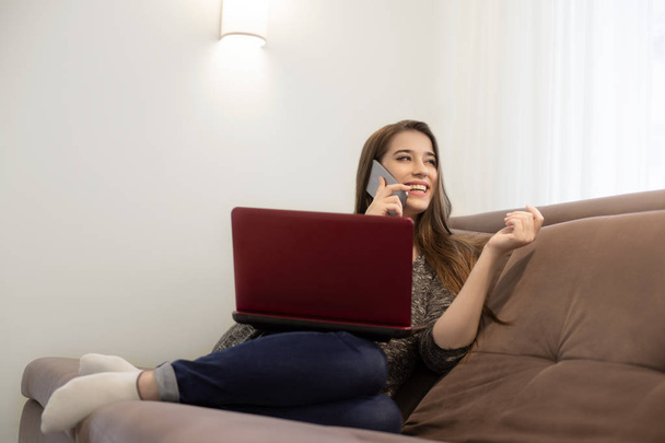 young beautiful smiling brunette girl sitting on the sofa talking on the phone and watching movie on laptop - Foto, Imagem