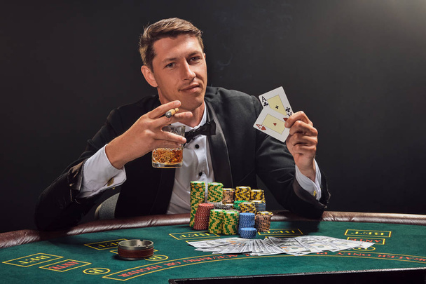 Bonito homem emocional está jogando poker sentado à mesa no cassino contra um holofote branco
. - Foto, Imagem
