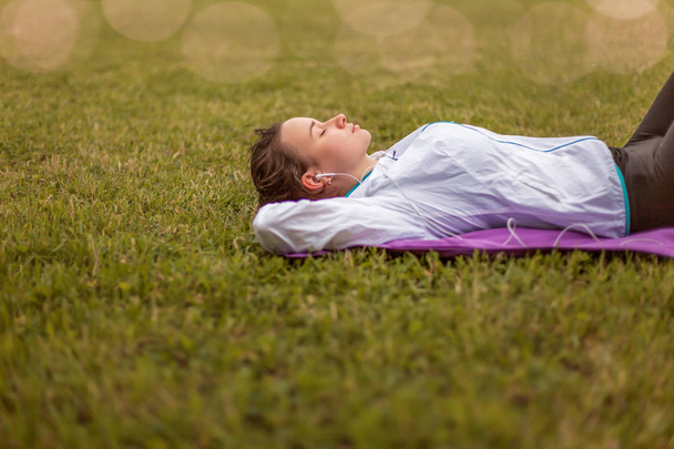 girl in sportswear resting lying on the grass, listening to music after training, jogging, exercising outdoors, urban style, healthy lifestyle - Photo, image