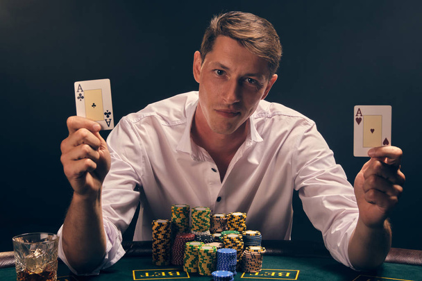 Schöner emotionaler Mann pokert am Tisch im Casino vor weißem Scheinwerferlicht. - Foto, Bild