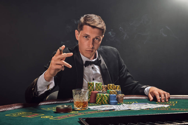 Knappe emotionele man speelt poker zitten aan de tafel in casino tegen een witte schijnwerper. - Foto, afbeelding