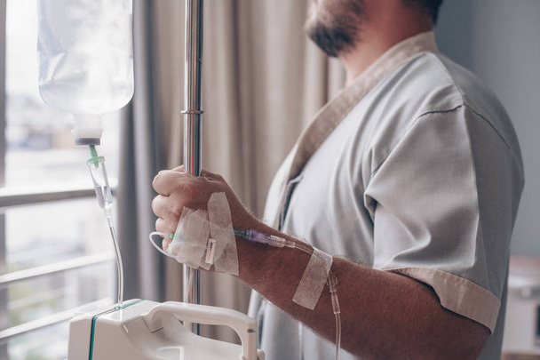 Ένας άντρας στέκεται σε ένα νοσοκομειακό θάλαμο και κοιτάζει από το παράθυρο. Σταγονόμετρο σε ένα χέρι Man σε ένα νοσοκομείο. - Φωτογραφία, εικόνα