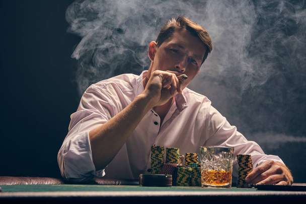 Όμορφος συναισθηματικός άντρας παίζει πόκερ καθισμένος στο τραπέζι του καζίνο ενάντια σε έναν λευκό προβολέα.. - Φωτογραφία, εικόνα