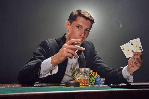 Handsome emotional man is playing poker sitting at the table in casino against a white spotlight. - Фото, зображення