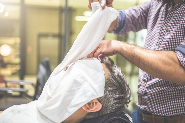 Peluquero aplicando una toalla caliente en la cara de un hombre antes de afeitarse en la barbería
. - Foto, imagen
