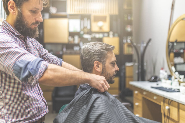 Professional barber ready to work and covering with cape bearded man on barbershop background. - Foto, afbeelding