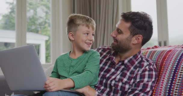 Front view of Caucasian father and son using laptop on sofa in a comfortable home. They are smiling and talking 4k - Filmagem, Vídeo