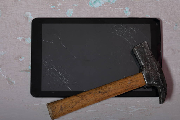 Tablet with a broken screen lies on the surface with a shabby paint. Nearby is a hammer. - Photo, image