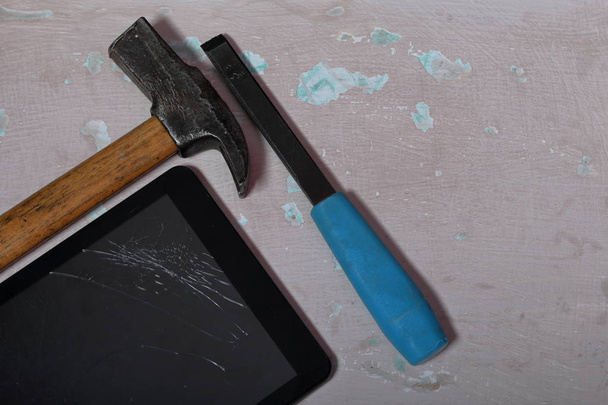 Tablet with a broken screen lies on the surface with a shabby paint. Nearby is a hammer and chisel. - Foto, Imagem