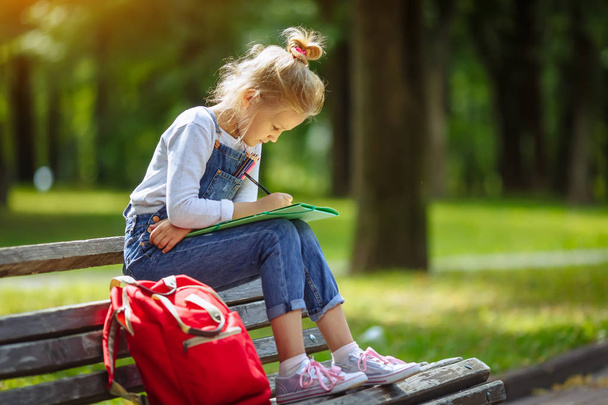 Pieni kaunis koulu tyttö piirustus värillisiä kyniä, istuu penkillä aurinkoisessa puistossa
 - Valokuva, kuva