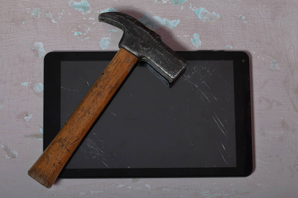 Tablet with a broken screen lies on the surface with a shabby paint. Nearby is a hammer. - 写真・画像