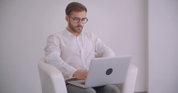 Closeup portrait of adult handsome bearded caucasian businessman in glasses using the laptop sitting in the armchair in the white office indoors - Záběry, video