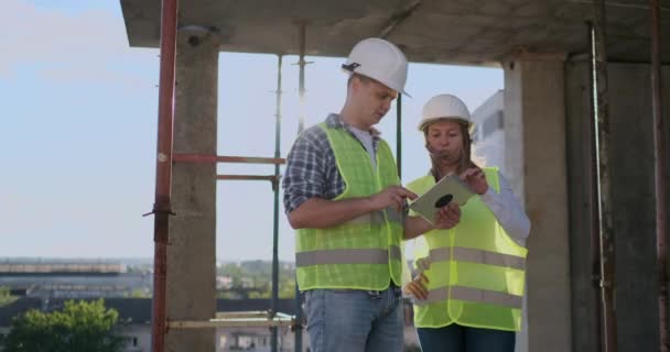 白いヘルメットとシャツで議論するタブレットコンピュータで建物の屋根の上に立っているエンジニアビルダーの男女. - 映像、動画