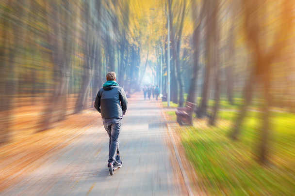 Giovane adulto cavalcando moderno scooter elettrico lungo bellissimo colorato parco cittadino autunnale. Uomo guida veicolo gadget attraverso multicolore valle degli alberi caduta. Sfocatura movimento
 - Foto, immagini