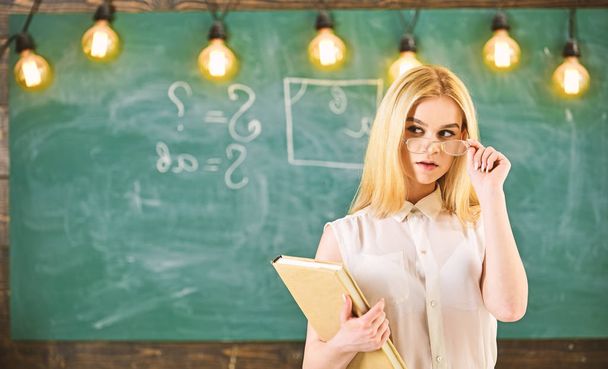 Woman with book starts lesson, gazes at audience while taking off eyeglasses. Teacher looks confident in eyeglasses, stand in classroom, chalkboard on background. Attractive lecturer concept - Photo, Image