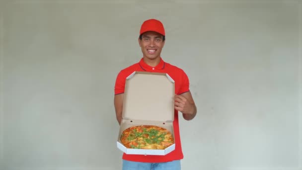 Knappe jonge levering werknemer in het rood uniform houdt een pizza, camera kijken en glimlachend, op grijze achtergrond - Video