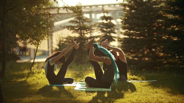zwei junge sportliche Frauen machen Yoga-Übungen mit einem Trainer im Park, umgeben von leuchtend grünen Bäumen - Foto, Bild