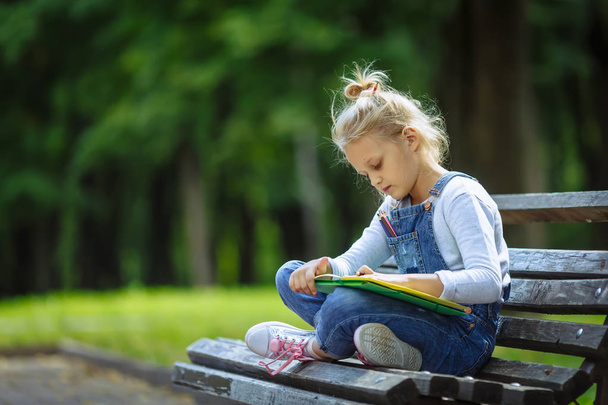 Pequeña niña hermosa escuela dibujo con lápices de colores, sentado en un banco en el parque soleado
 - Foto, Imagen