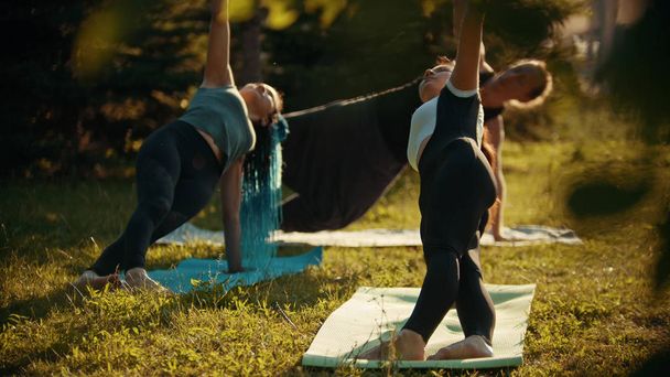 Dos mujeres atléticas jóvenes y un hombre haciendo ejercicios de yoga en el parque rodeado de árboles verdes brillantes
 - Foto, imagen