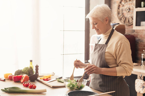 Positiivinen vanhempi nainen ruoanlaitto tuoretta salaattia keittiössä
 - Valokuva, kuva