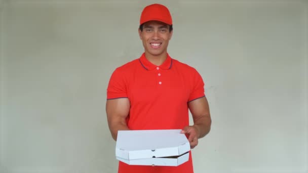 Handsome young delivery worker in red uniform is holding a pizza, looking at camera and smiling, on gray background - Footage, Video