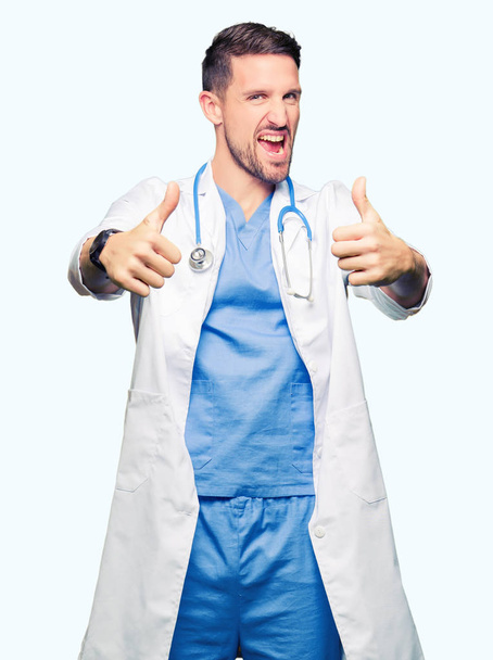 Handsome doctor man wearing medical uniform over isolated background approving doing positive gesture with hand, thumbs up smiling and happy for success. Looking at the camera, winner gesture. - Fotografie, Obrázek