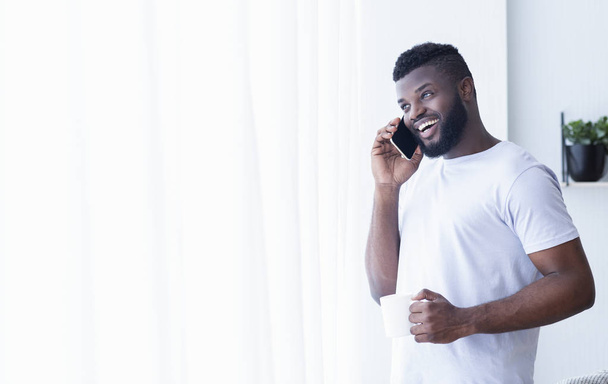 Hombre negro alegre hablando en el teléfono móvil, mirando a la ventana con café
 - Foto, Imagen