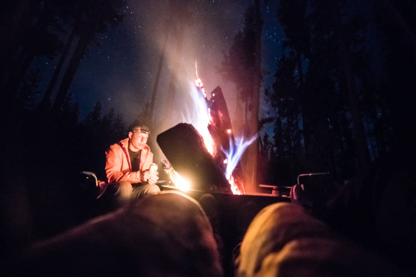 People enjoy the campfire at night sitting drinking and relaxing at campsite - 写真・画像