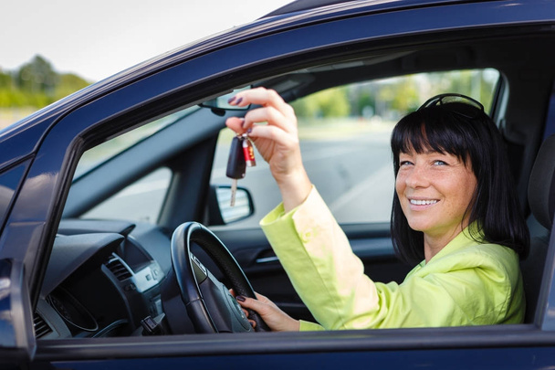 春夏の気分で彼女の車の運転席に座って手に車のキーを持つ幸せな笑顔ブルネットの女性35歳。女性運転。頭にサングラスをかけたグリーンジャケットを着た - 写真・画像
