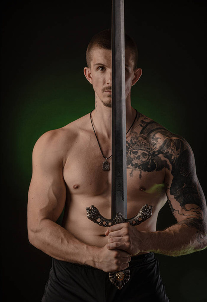 muskularny facet z gołymi tułowia stwarzających na ciemnym tle z mieczem - Zdjęcie, obraz