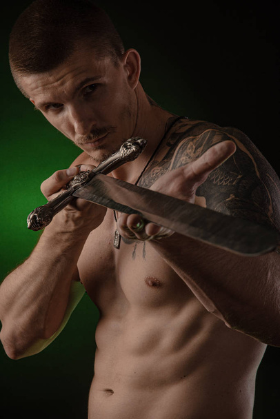 Мускулистый парень с голой туловищем позирует на темном фоне с мечом
 - Фото, изображение