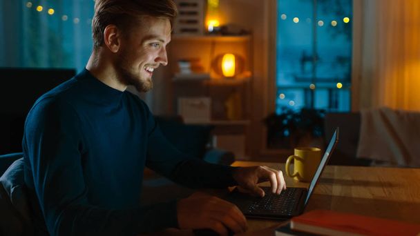 Evening at Home: Portrait of Handsome Man Sitting at His Desk Working on a Laptop. Smiling Freelancer Working on Computer. Side View Shot. - Foto, Imagem