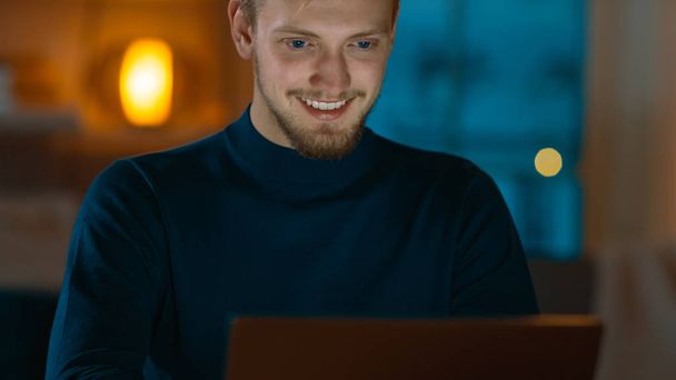 Evening at Home: Portrait of Handsome Man Sitting at His Desk Working on a Laptop. Smiling Freelancer Working on Computer. - 写真・画像