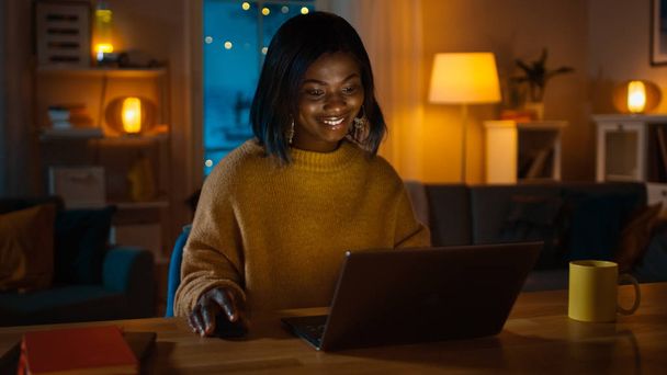 Portrait of Beautiful Smiling African American Girl Using Laptop while Sitting at Her Desk at Home. In the Evening Creative Woman Works on a Computer In Her Cozy Living Room. - Фото, изображение