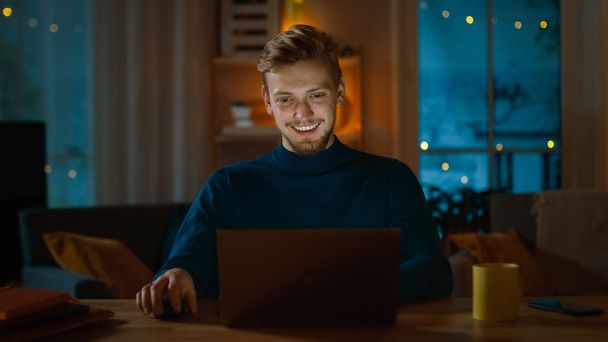 Handsome Smiling Man Works on a Laptop while Sitting at His Desk at Home. Portrait of a Young Freelancer Works on Computer in His Cozy Living Room with Warm Evening Lighting in Background. - Foto, afbeelding