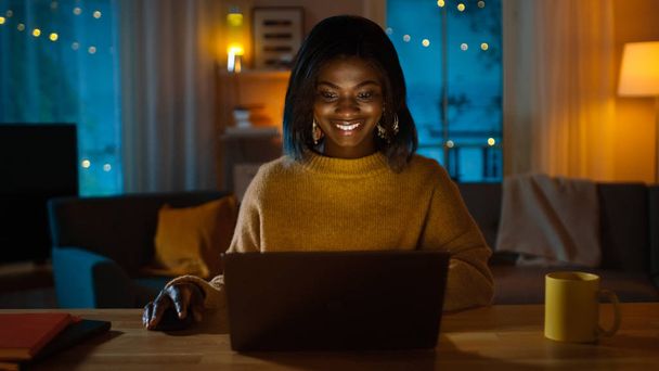 Portrait of Beautiful Smiling Black Girl Using Laptop while Sitting at Her Desk at Home. In the Evening Creative Woman Works on a Computer In Her Cozy Living Room. - Zdjęcie, obraz