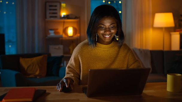 Portrait of Beautiful Smiling African American Girl Using Laptop while Sitting at Her Desk at Home. In the Evening Creative Woman Works on a Computer In Her Cozy Living Room. - Fotografie, Obrázek