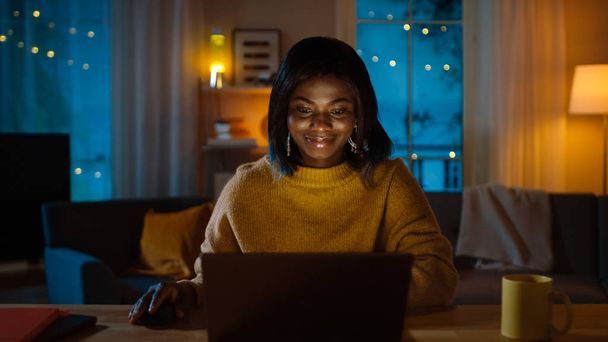 Portrait of Beautiful Smiling Black Girl Working on a Laptop while Sitting at Her Desk at Home. In the Evening Creative Woman Works on a Computer In Her Cozy Living Room. - Фото, изображение