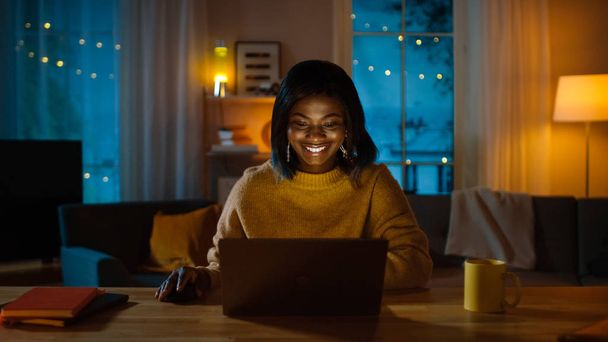 Retrato de una hermosa chica negra sonriente trabajando en una computadora portátil mientras está sentada en su escritorio en casa. Por la noche, la mujer creativa trabaja en una computadora en su acogedora sala de estar
. - Foto, imagen