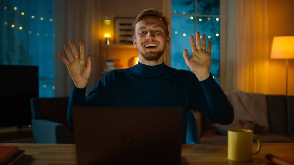 Illalla: Onnellinen nuori mies tanssii istuessaan työpöydällään, Hän käyttää kannettavaa tietokonetta ja kuuntelee musiikkia
. - Valokuva, kuva