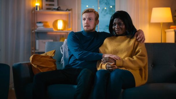 Happy Young Couple Watching Horror Movie on TV while Sitting on a Couch, they got Scared, Girl Covers eyes. Handsome Caucasian Boy and Black Girl in Love Spending Time Together. - Zdjęcie, obraz