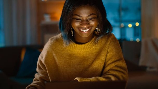 Portrait of Beautiful Black Girl Uses Laptop while Sitting at Her Desk at Home, Her Face is Illuminated with a Screen Light. In the Evening Creative Woman Works on a Device In Cozy Living Room. - Foto, Imagen