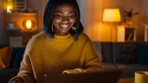 Portrait of Beautiful Black Girl Uses Laptop while Sitting at Her Desk at Home, Her Face is Illuminated with a Screen Light. In the Evening Creative Woman Works on a Device In Cozy Living Room. - Photo, image